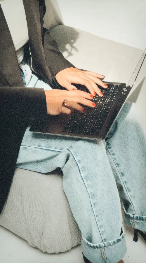 A woman in a blazer and denim typing on a laptop balanced on her lap in a bright studio.